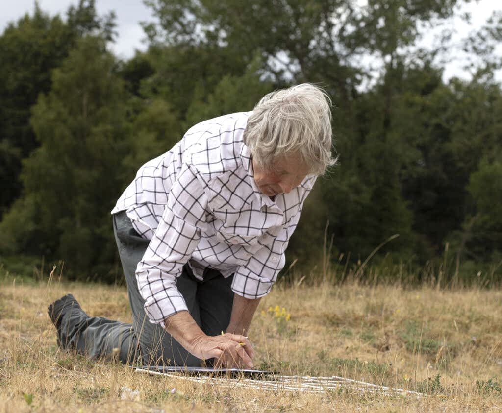 A woman wearing a white and black checked shirt is kneeling on the ground investigating a square on the ground