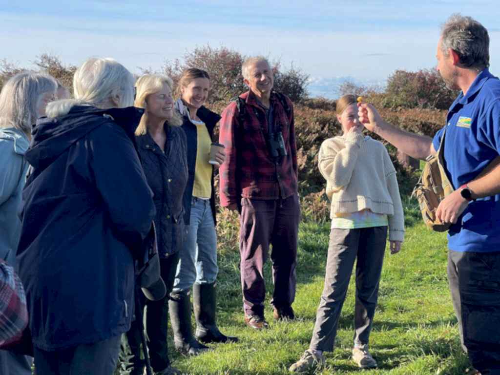 A group of people stood in a semi circle in a field watching and listening to a man in a blue tshirt. The man is holding a mushroom.