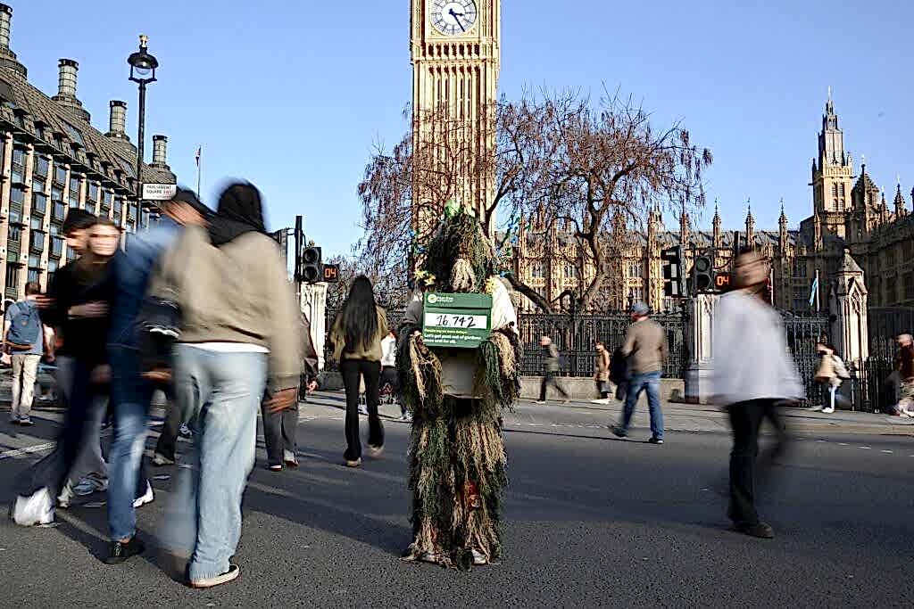 Someone wearing a bog costume holding a box showing 16,000 have signed a petition in front of Parliament