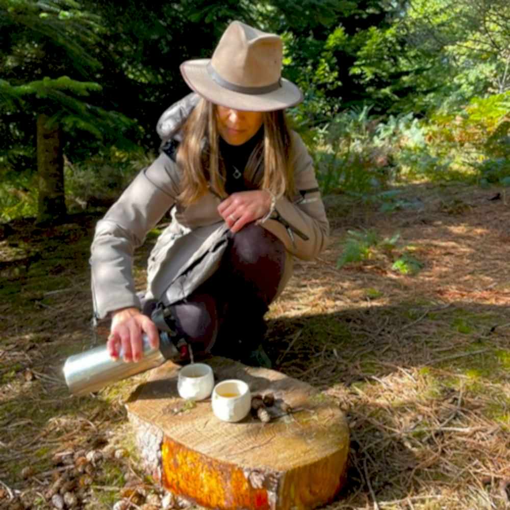 Photograph shows a woman crouching on the floor, she is pouring tea from a flask into two cups resting on a wooden stump.