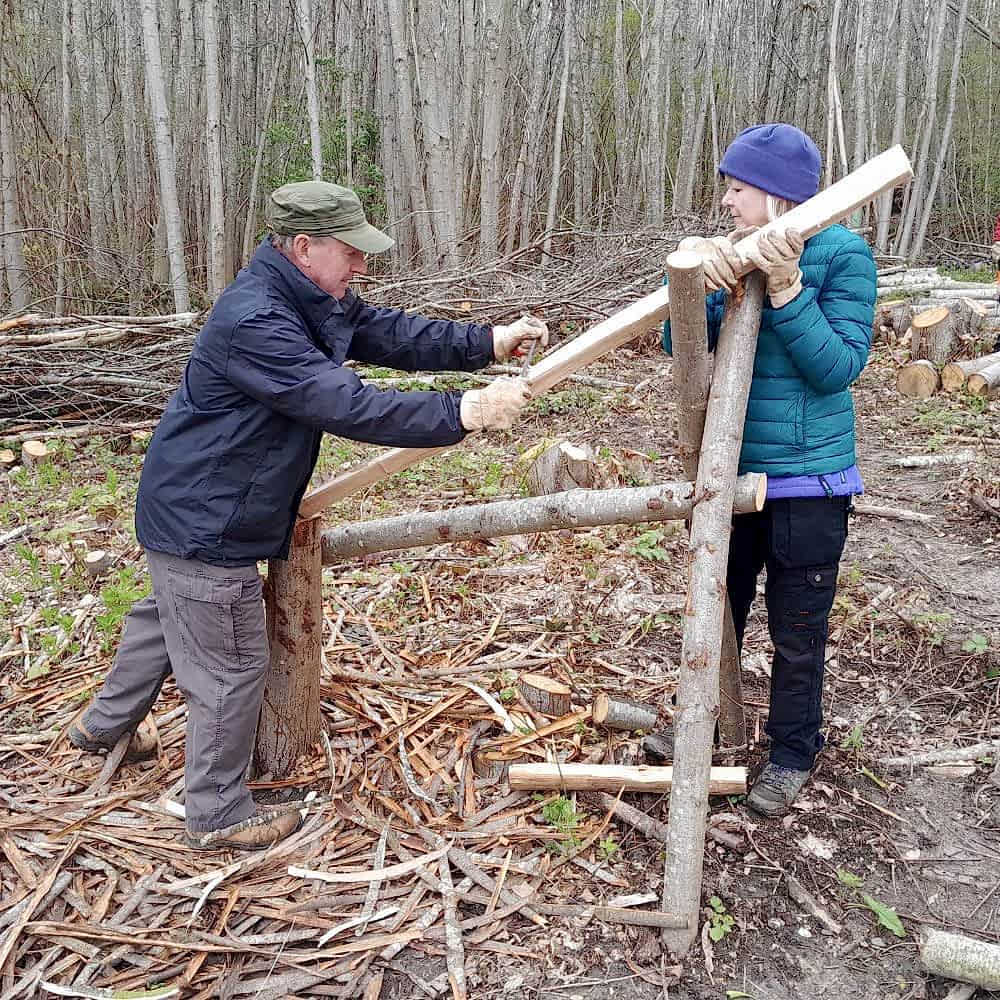 Photograph shows two people, both volunteers, wood working in the middle of a clearing in a forest.
